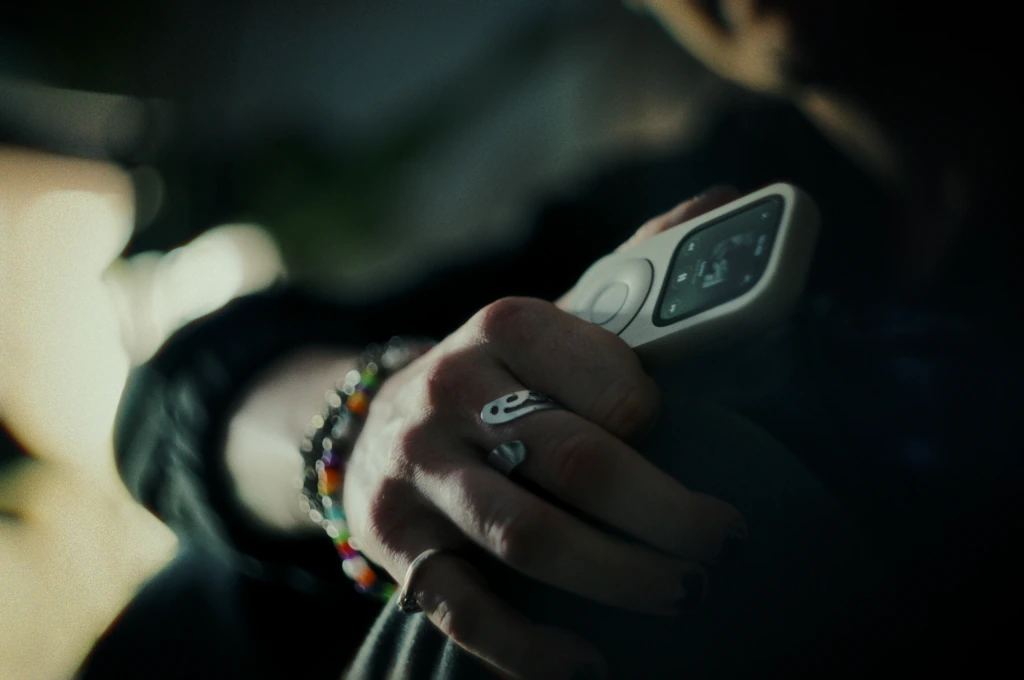 Una mano con anillos sostiene un RePod plateado con controles de música visibles, en un interior con luz suave.